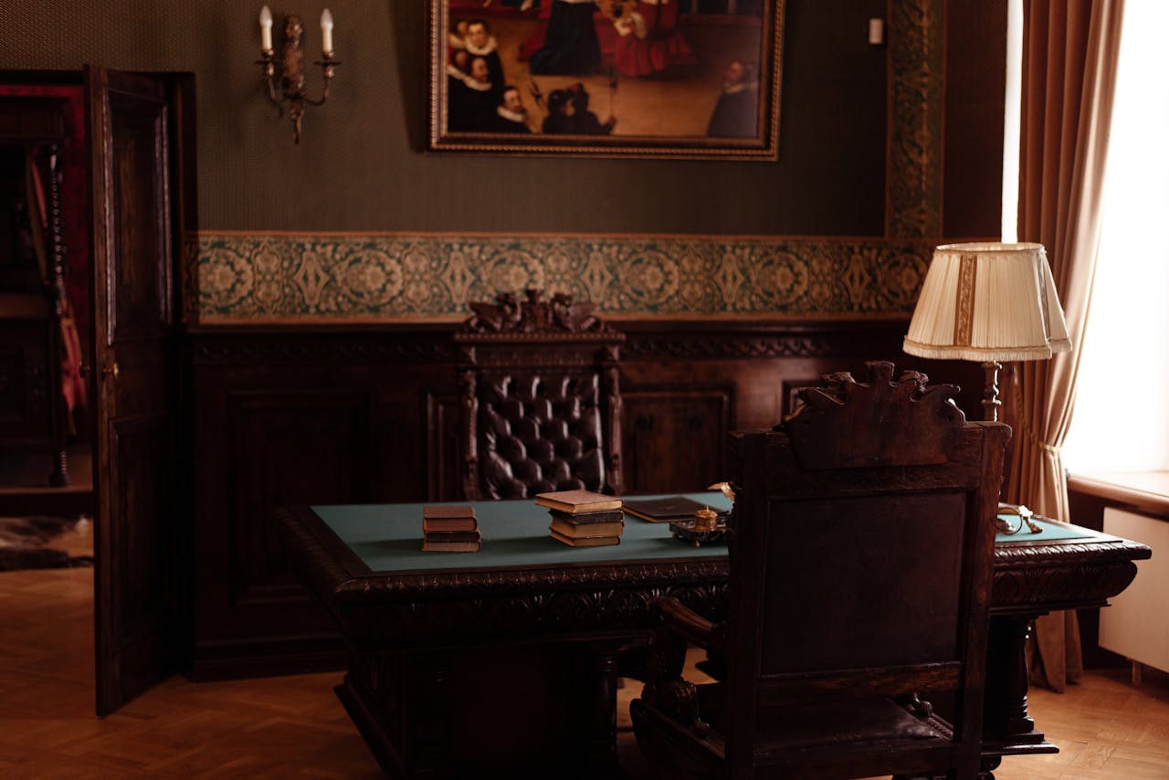 gallery-3 An ornate study room featuring antique wooden furniture, books, and a classic lamp.